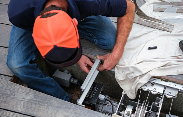 A person in a red and black cap and blue shirt installs or repairs mechanical equipment under a deck, using tools and metal parts, with a white cloth covering other components nearby.